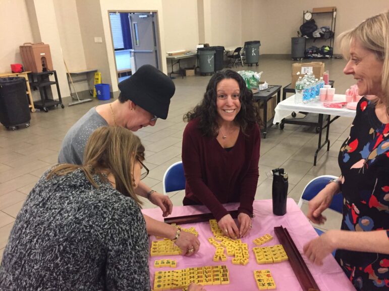 Four women sit around a table covered with yellow mahjong tiles, arranging the pieces. The background shows folding tables with water bottles and other supplies in a large indoor room.