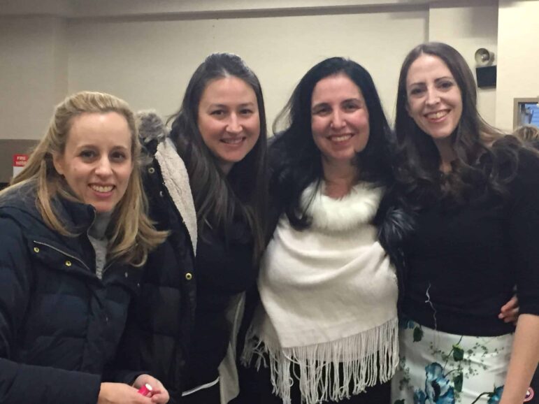 Four women are standing closely together indoors, smiling at the camera. One woman is wearing a black jacket, and the others are dressed in dark tops, with one wearing a white shawl. They appear to be at a social gathering.