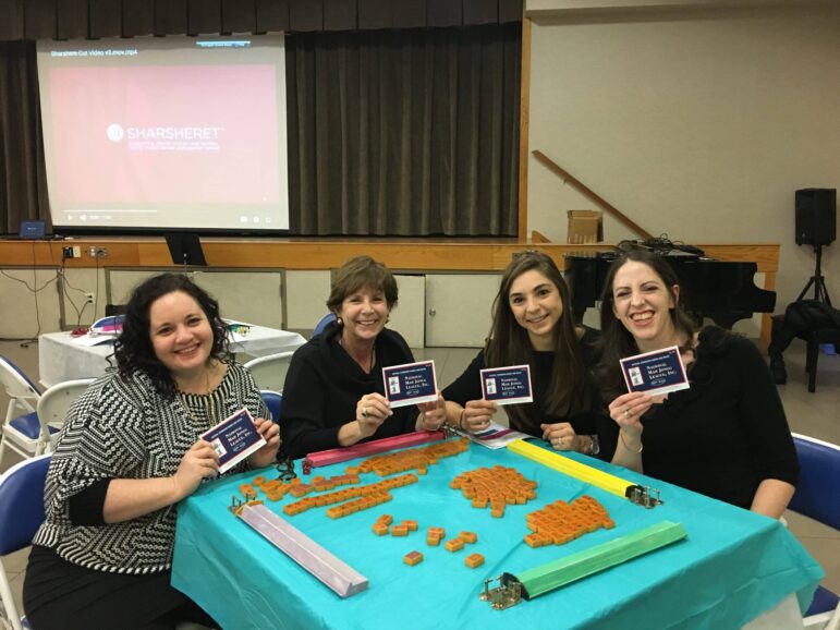 Four women sit at a table covered with orange cookie cutters, decorated cookies, and colored icing bags. Each holds a card. A presentation screen and a piano are visible in the background.