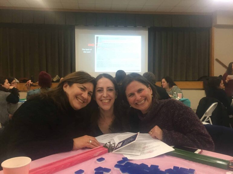 Three women sit together at a table covered with papers, folders, and blue game pieces, smiling at the camera. More people are seated in the background, and a presentation is projected on a screen.