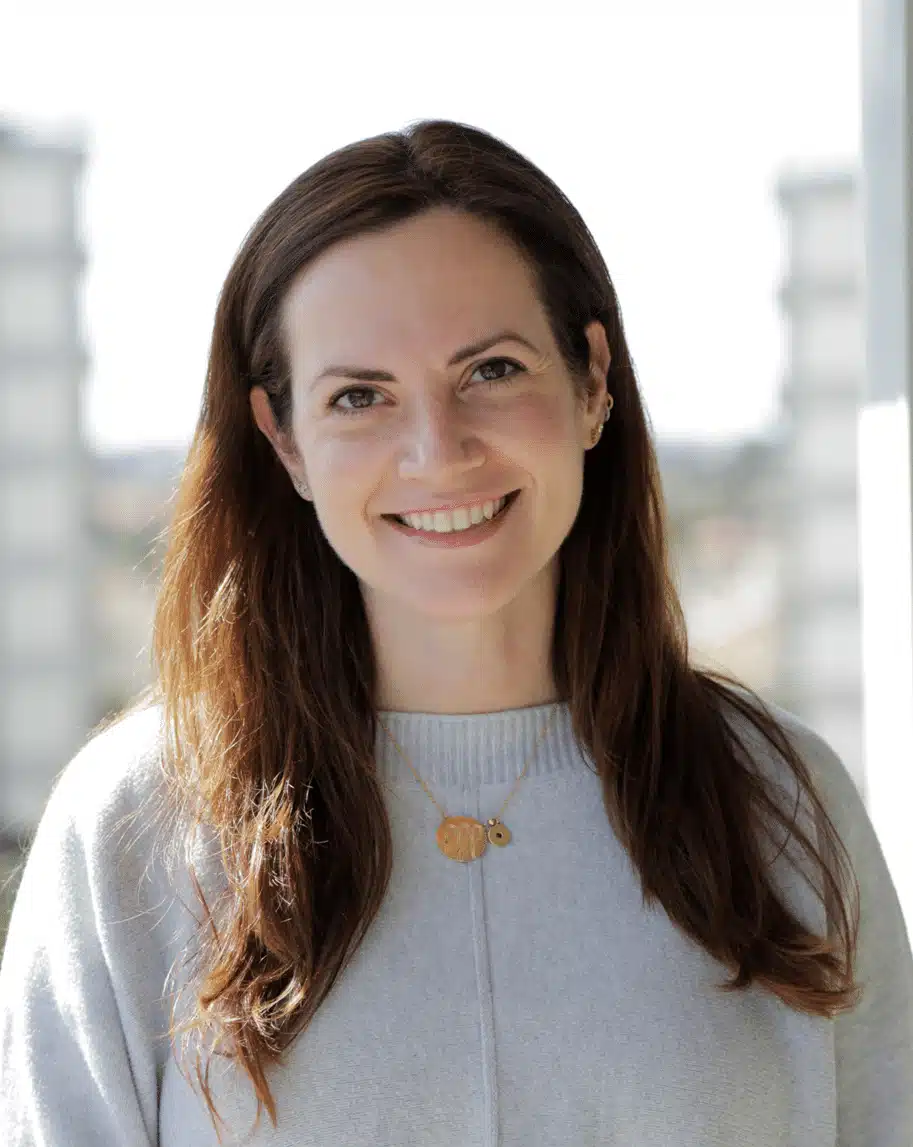 A woman with long brown hair smiles at the camera. She is wearing a light grey sweater and a gold necklace. The background is blurred with two tall structures visible on either side.