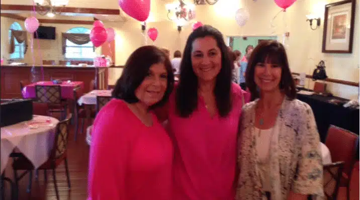 Three women stand together and smile for the camera in a decorated indoor space. Pink and white balloons are visible in the background, and the women wear pink and light-colored outfits. Tables and chairs are arranged around them.