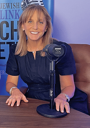 A woman with shoulder-length blonde hair wearing a navy blue dress sits at a table with a microphone in front of her. A blurred blue banner with partial text is in the background.