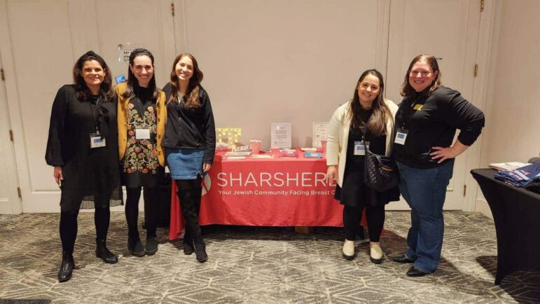 Five women stand and smile in front of a red table with a “Sharsheret” banner, informational materials, and display items; the setting appears to be an indoor event or conference.