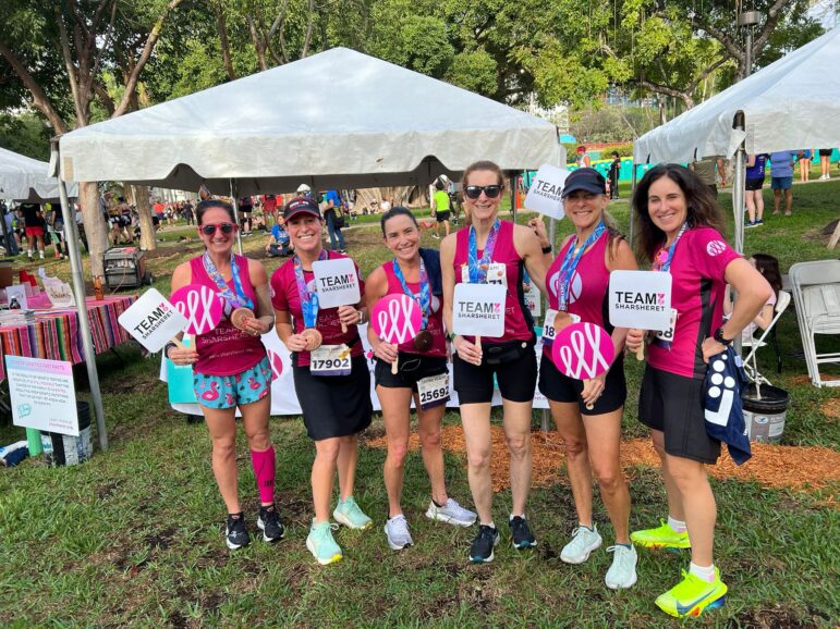 Six women in athletic wear stand on grass, holding medals and “TEAM? #BeMoreThanPink” signs, smiling at the camera. Tents and other people are visible in the background, suggesting a race event.