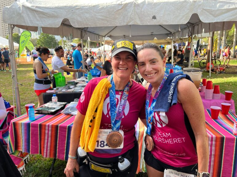 Two women wearing running gear, finisher medals, and race bibs smile at the camera in front of a tent. Other runners and volunteers are visible in the background at the outdoor event.