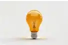 A light bulb filled with an orange liquid stands upright on a plain, light-colored surface, with a subtle reflection and shadow beneath it.