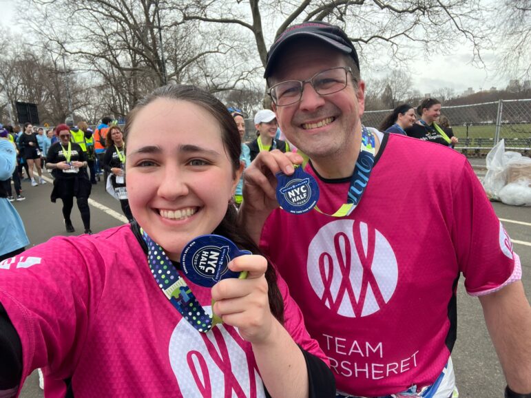 Two runners wearing pink Team Sharsheret shirts smile and hold up NYC Half Marathon medals. Other participants can be seen in the background on an overcast day.