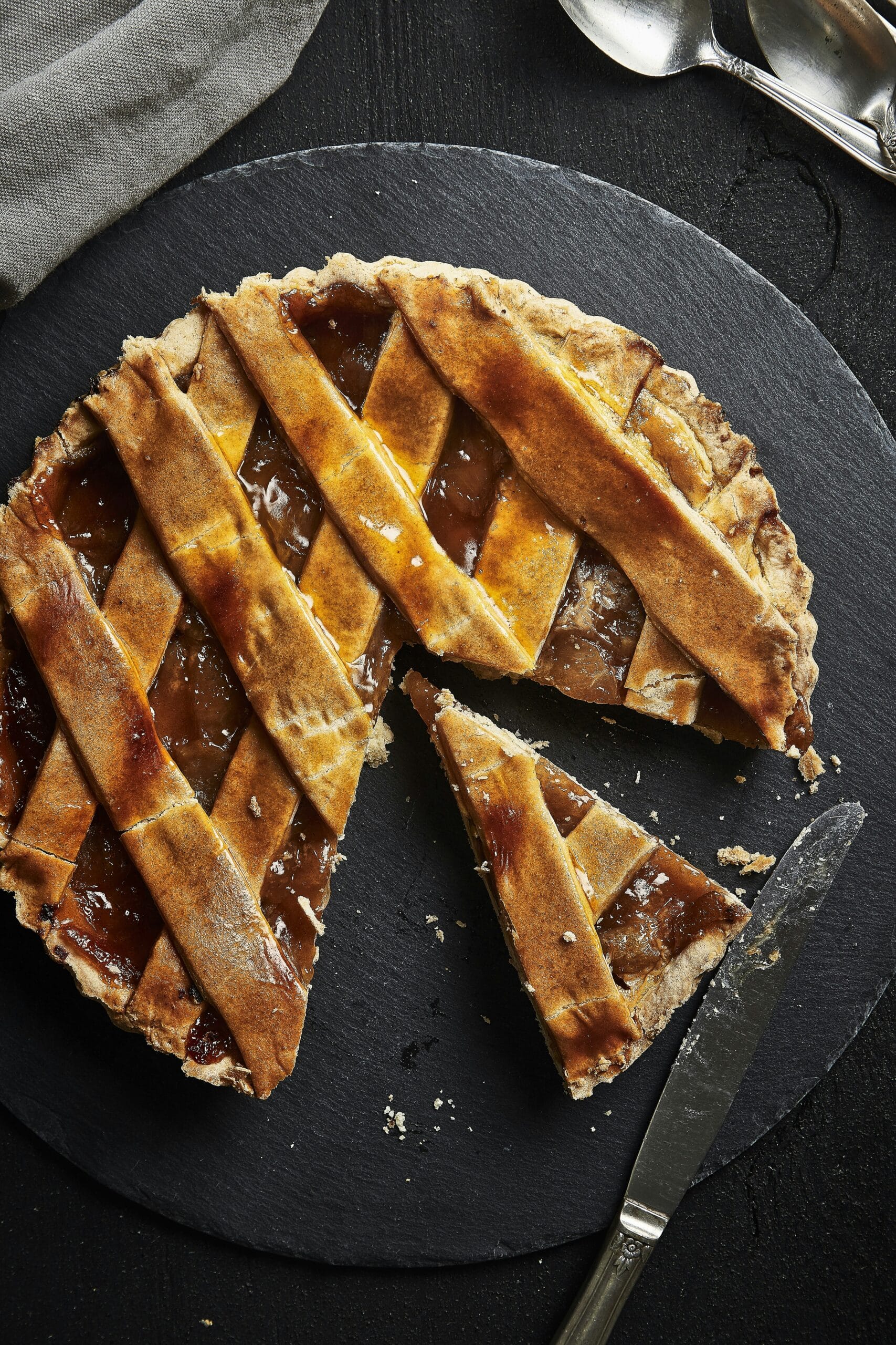 A round lattice-top pie sits on a dark slate serving board, with one slice removed and placed slightly away from the rest. A knife rests nearby, and two spoons are partially visible in the corner.