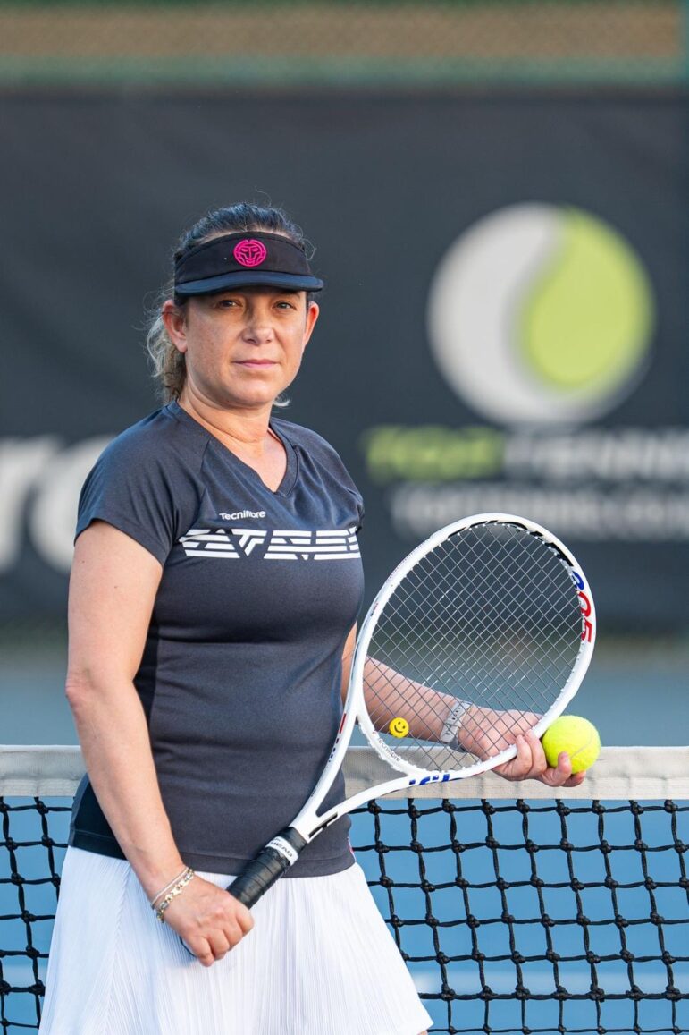 A woman in a black visor and dark shirt stands on a tennis court, holding a tennis racket and a yellow tennis ball, embodying the sense of emotional and spiritual healing that outdoor sports can bring; behind her, a net and a blurred logo on a banner are visible in the background.