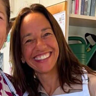 A woman wearing a white apron with a pink logo stands in a kitchen, smiling at the camera, radiating warmth and positivity—a space that feels infused with emotional and spiritual healing. Shelves with books and a green watering can are visible in the background, suggesting a nurturing environment. Another person is partly visible on the left.