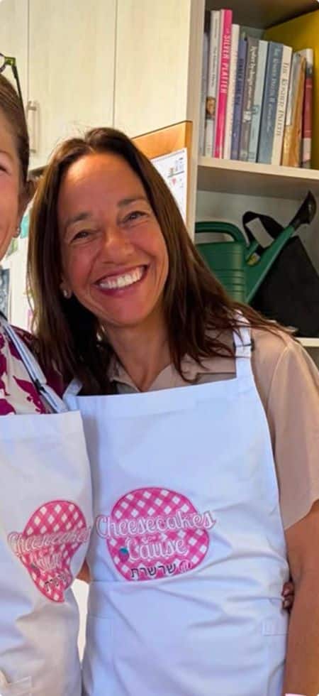 A woman wearing a white apron with a pink logo stands in a kitchen, smiling at the camera, radiating warmth and positivity—a space that feels infused with emotional and spiritual healing. Shelves with books and a green watering can are visible in the background, suggesting a nurturing environment. Another person is partly visible on the left.