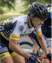 A cyclist wearing a helmet, sunglasses, and a white and navy cycling jersey rides a bike during a race, with another cyclist partially visible in the background.