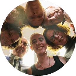 Six adults stand in a circle, looking down toward the camera and smiling. The photo is taken from below, creating a circular composition with their faces around the edge.