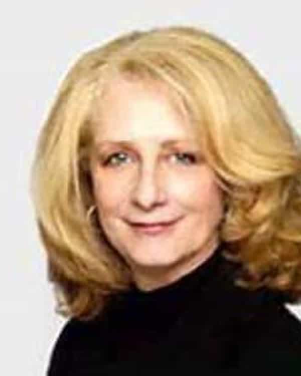 A woman with shoulder-length blonde hair wearing a black top poses in front of a plain, light-colored background. She is facing the camera and has a neutral expression.