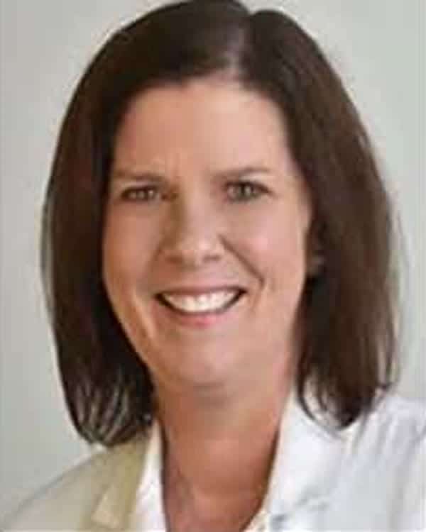 A woman with straight, shoulder-length brown hair smiles at the camera. She is wearing a white collared shirt and is posed in front of a plain, light-colored background.