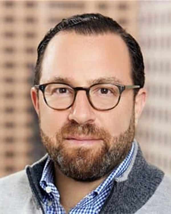 A man with short dark hair, a beard, and glasses is wearing a light blue checkered shirt and a grey zip-up sweater. The background is blurred with a pattern of light brown and dark brown squares.