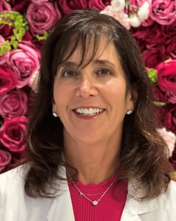 A woman with long dark hair wearing a white lab coat and pink top stands in front of a wall covered with pink and red roses. She is smiling and facing the camera.