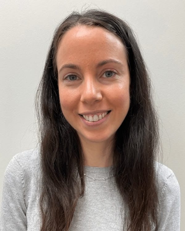 A person with long brown hair wearing a short-sleeved light grey top stands in front of a plain light grey background, smiling at the camera.