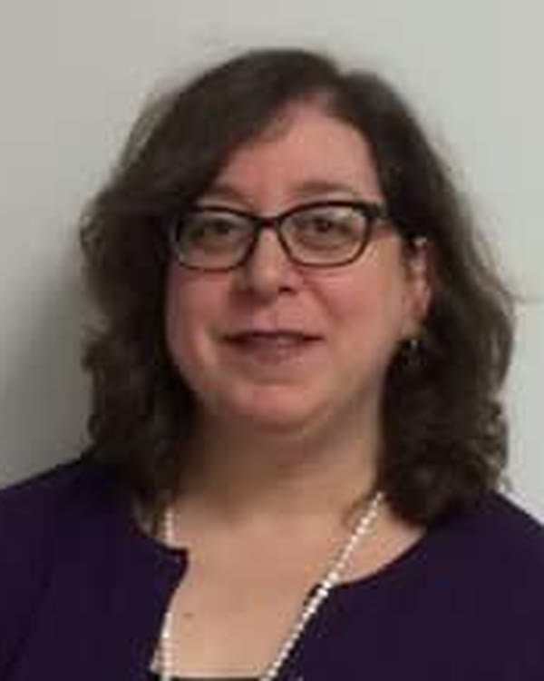 A woman with shoulder-length brown hair and glasses is wearing a purple cardigan over a black top and a long pearl necklace, standing against a plain light-colored background.
