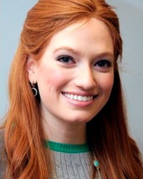 A woman with long red hair, fair skin, and a wide smile is facing the camera. She is wearing a gray sweater with a green collar and small hoop earrings. The background is plain and light-colored.