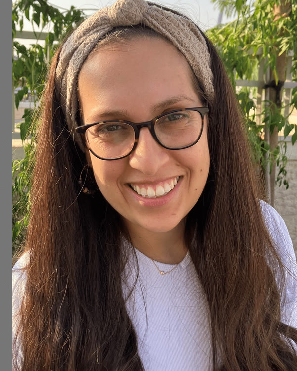 A woman with long brown hair, glasses, and a beige headband is smiling at the camera. She is wearing a white top and standing outdoors with greenery in the background.