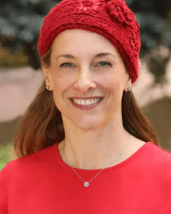 A woman with long brown hair wears a red knit hat and a red top, smiling at the camera outdoors. She is also wearing a necklace with a small round pendant. The background is blurred.