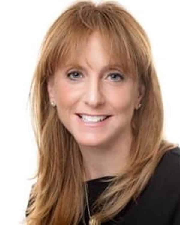A woman with straight, light brown hair and bangs, wearing a black top and a gold necklace, smiles at the camera against a white background.