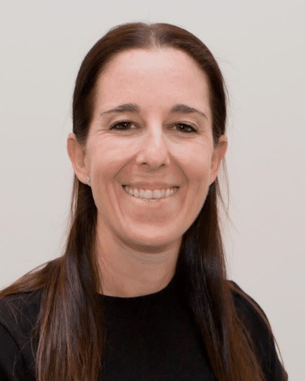 A woman with straight, long brown hair and a black short-sleeve top is smiling at the camera. The background is plain light grey with grey bars on both sides of the image.