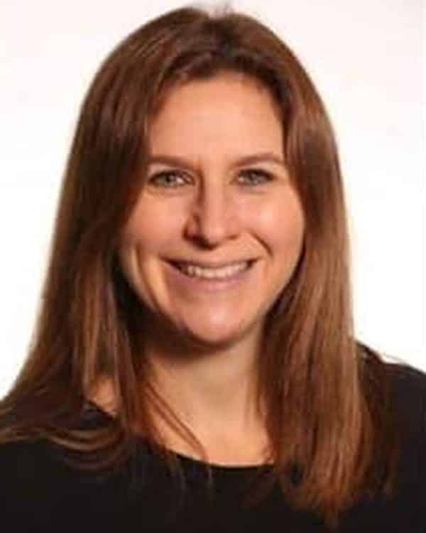 A woman with long brown hair and a black top smiles at the camera against a plain white background.