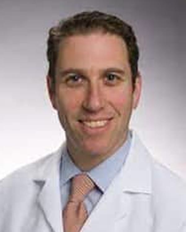 A man wearing a white medical coat, light blue shirt, and light-colored tie, smiling at the camera against a neutral background.