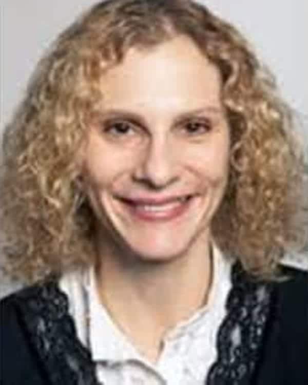 A person with curly blonde hair is smiling at the camera. They are wearing a white collared shirt with a black garment that has lace trim. The background is plain and light-colored.