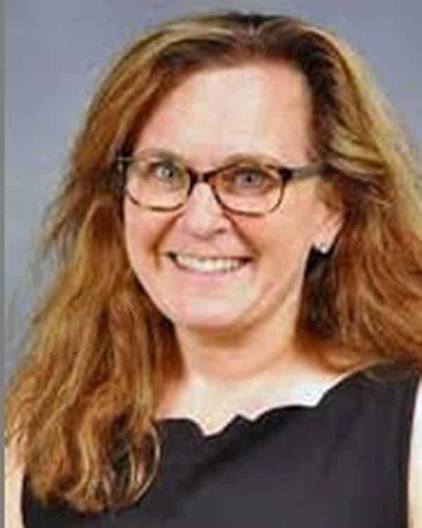 A woman with long, light brown hair and glasses smiles at the camera. She is wearing a sleeveless black top and is photographed against a plain gray background.