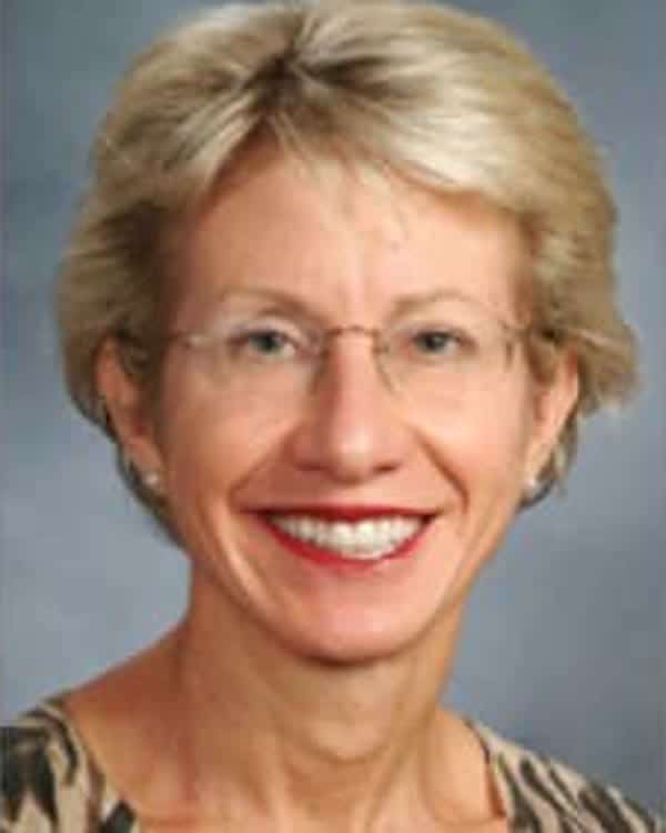 A middle-aged woman with short, light blonde hair and glasses, smiling at the camera. She is wearing a patterned top and small earrings, shown against a plain blue-gray background.