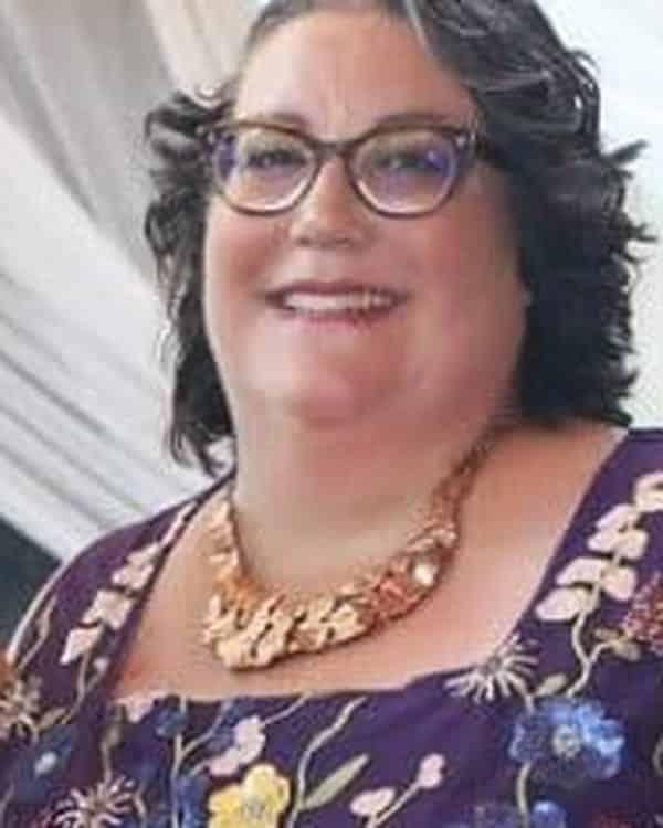 A woman with curly dark hair and glasses smiles at the camera. She is wearing a purple dress with colorful floral embroidery and a gold necklace. White drapery is visible in the background.