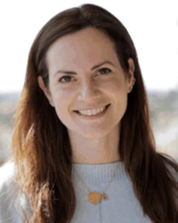A woman with long brown hair wearing a light grey sweater and a gold necklace is smiling at the camera. The background is blurred with two white structures visible on either side.
