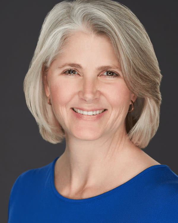 A person with gray, shoulder-length hair is smiling at the camera. They are wearing a blue top and are posed in front of a plain, dark background.