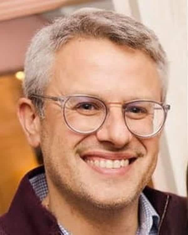 A man with short gray hair and glasses smiles at the camera. He is wearing a maroon jacket over a blue and white checkered shirt. The background is softly blurred in warm tones.