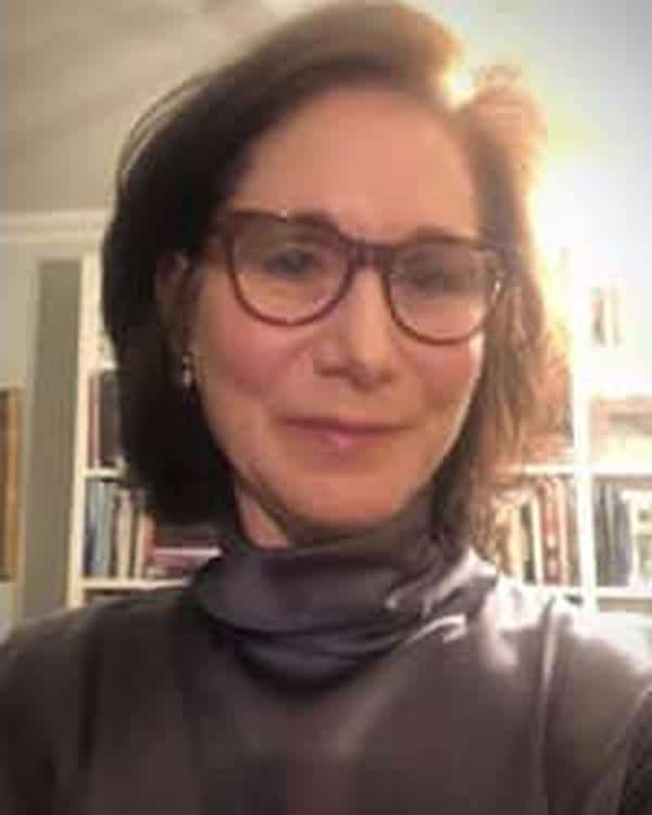 A woman with short brown hair and glasses is wearing a high-necked top and is standing indoors in front of a bookcase, facing the camera and smiling slightly. The lighting is warm and bright.