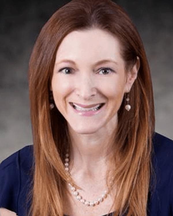 A woman with straight, long red hair, wearing a navy blue blouse and a pearl necklace, smiles at the camera against a plain, dark gradient background.