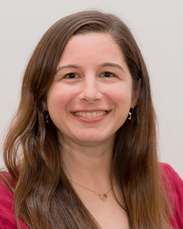 A woman with long brown hair wearing a short-sleeved, dark pink top stands in front of a plain light background, smiling at the camera.