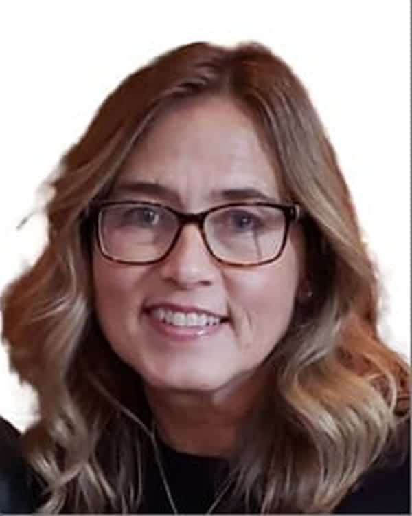 A woman with wavy, shoulder-length blonde hair and glasses is smiling at the camera. She is wearing a dark top and is posed against a plain white background.
