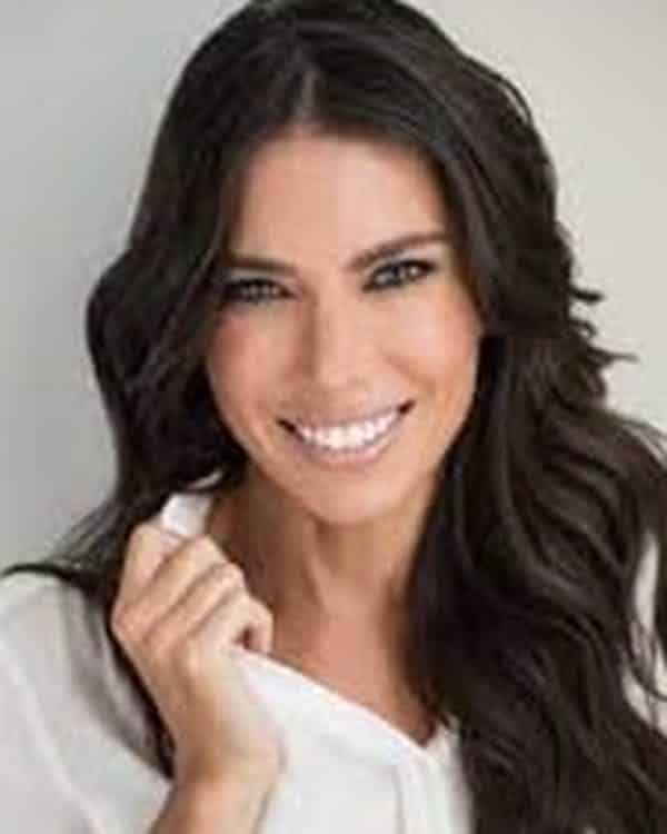 A woman with long, dark wavy hair is smiling at the camera. She is wearing a white blouse and sitting against a plain light background. Her right hand is touching her hair.