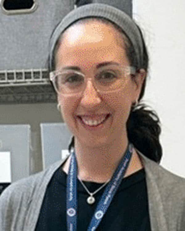 A woman wearing glasses, a gray sweater, and a work badge stands behind a table with books, folders, yarn, and small bags. Office shelves and supplies are visible in the background.