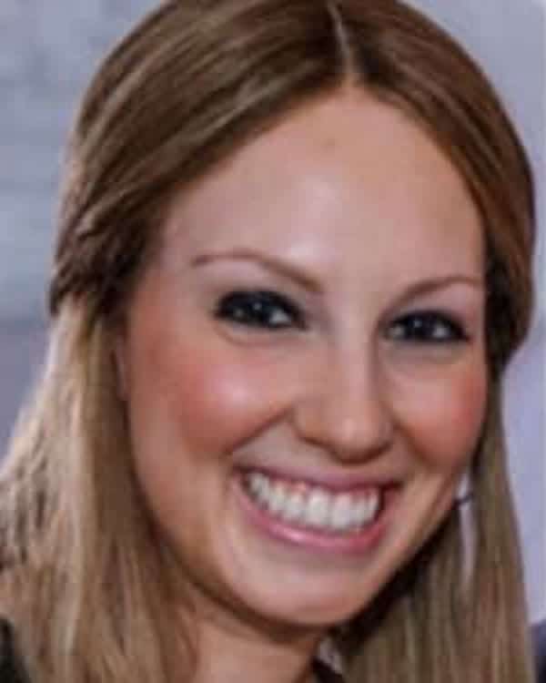A woman with long light brown hair is smiling widely at the camera. She is indoors, and the background is blurred.