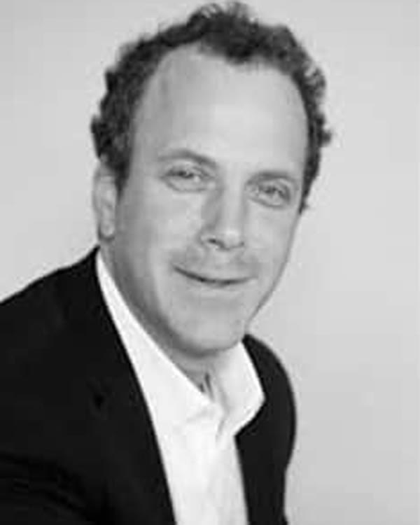 A man with short, curly hair is wearing a dark suit jacket and a white shirt. He is facing the camera and smiling slightly. The background is plain and light-colored. The image is in black and white.