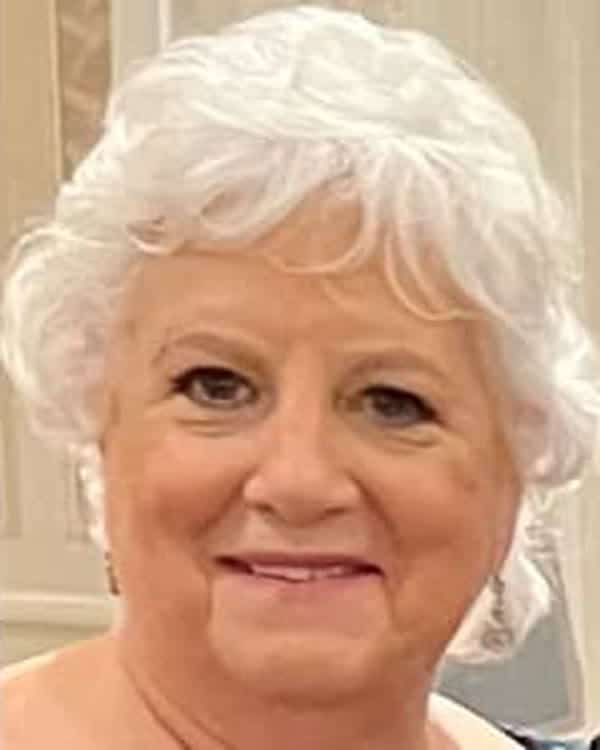 An older woman with short, curly white hair is smiling slightly. She is wearing light makeup, and the background appears to be indoors with neutral-colored walls.