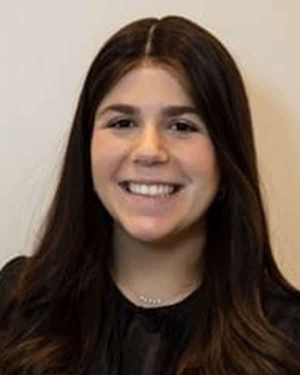 A person with long, dark brown hair is smiling at the camera. They are wearing a black top and are posed in front of a plain light-colored background.