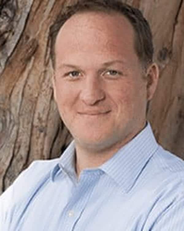 A man with short light brown hair wearing a light blue button-up shirt stands in front of a large tree trunk, looking at the camera with a slight smile and arms crossed.