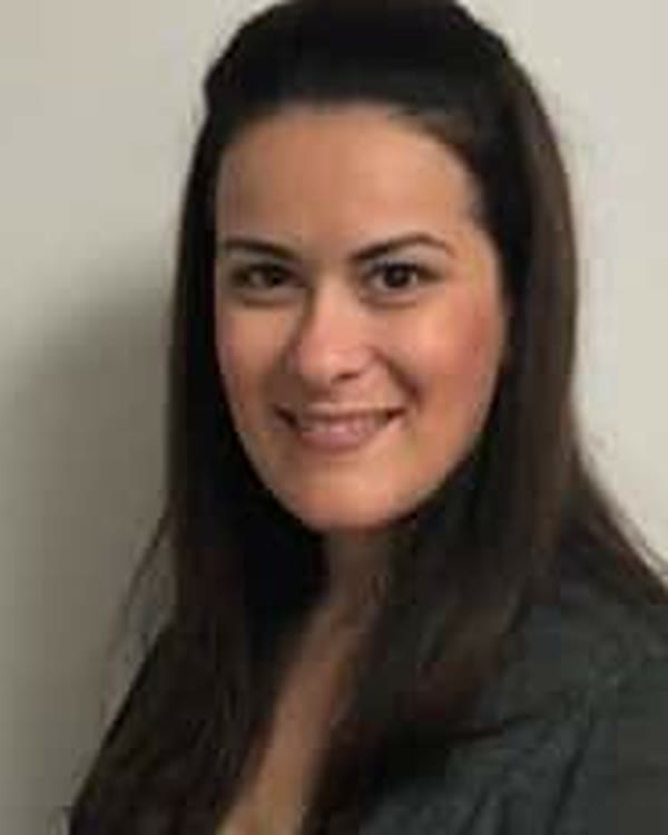 A woman with long dark hair, wearing a dark blazer over a top, stands in front of a plain light-colored wall, smiling at the camera.